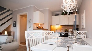 Un comedor con una mesa blanca dispuesta para cuatro, una araña y una cocina al fondo.