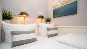 Una cama bien hecha con almohadas a rayas blancas y grises, dos lámparas de pared, una planta en maceta y un cuadro de un faro en la pared.
