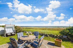 En udendørs terrasse med et rundt træbord og stribede stole, med udsigt over et kystlandskab med en stenvæg og havudsigt.