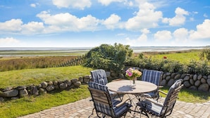 A patio with a round table and chairs, a vase of flowers, and a view of a coastal landscape. - Am Watt 1 - Schöne Erdgeschoßwohnung auf Einer Ebene, ca. 130 m² Groß, für bis zu 4 Personen (Sylt)