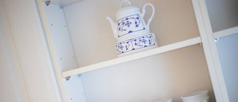 A white cabinet with two shelves. The top shelf holds a white teapot with blue floral designs. The bottom shelf has four white mugs with blue floral designs.