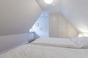 Un dormitorio con techo inclinado, paredes blancas y una cama blanca. Se observa un armario blanco y una puerta al fondo.