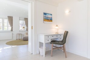 Ein helles Zimmer mit Schreibtisch, Stuhl und Lampe. An der Wand hängt ein Bild, und ein Fenster mit Vorhängen ist zu sehen.