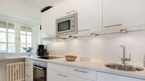 A modern kitchen with white cabinetry, a built-in microwave, a stainless steel sink, and a window with a view of greenery. - Weber EG - Weber EG - Wohnen Unter Reet im Kapitänsdorf Keitum (Sylt-Ost)