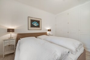 Un dormitorio con dos camas, una mesita de noche con lámpara, un cuadro enmarcado en la pared y un armario.