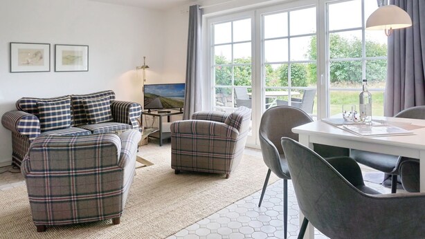 Una sala de estar moderna con un conjunto de sofá, una mesa de comedor con sillas, un televisor y ventanas grandes que ofrecen vistas al exterior.