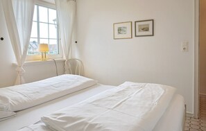 Quarto simples e limpo, com duas camas de solteiro, uma janela com cortinas e duas imagens emolduradas na parede.