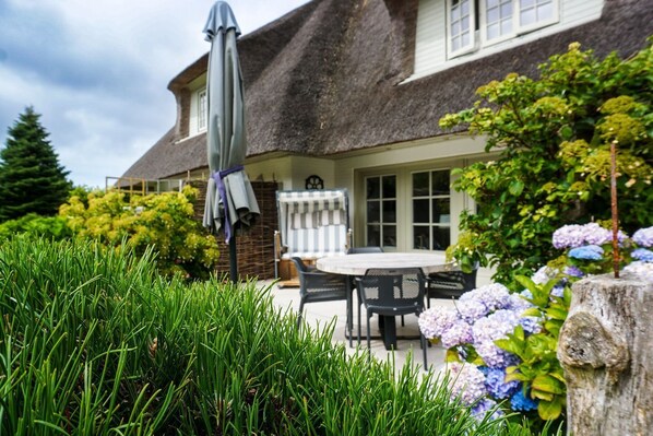 A house with a thatched roof, a dining area with a table and chairs, and a garden with hydrangeas.