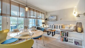 En moderne stue med spiseområde, store vinduer med persienner, en sofa, et sofabord og et bogskab.