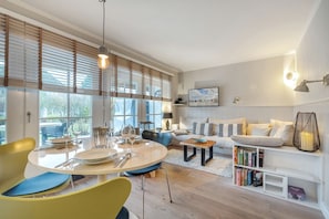 A modern living room with a dining area, large windows with blinds, a sofa set, a coffee table, and a bookshelf. - Lille Hygge - Modere, Komplett Ebenerdige Wohnung in Ruhiger Lage (Sylt-Ost)