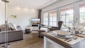 A modern living room with a sofa, a coffee table, a dining table set for two, and a TV. - Lüüvwai II - In Einer Reetgedeckten Wohnanlage in Südlage Nahe dem Benen-diken-hotel (Sylt-Ost)