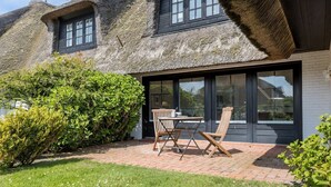 A house with a thatched roof, a large window, and a patio with a table and chairs. - Lüüvwai II - In Einer Reetgedeckten Wohnanlage in Südlage Nahe dem Benen-diken-hotel (Sylt-Ost)