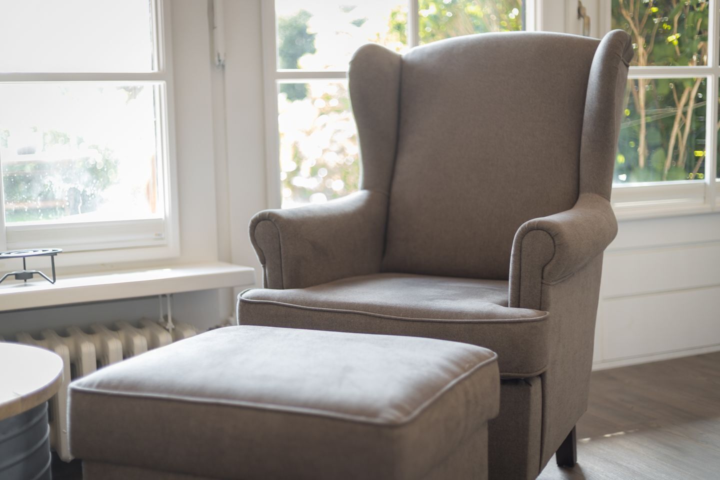 A grey armchair with a matching ottoman in a room with a large window, a radiator, and a small table.