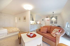 A cozy, well-lit room with a bed, a sofa, a small table with a cup, and a kitchen area in the background. - Haus Süderende - Gemütliches Appartement mit 34 qm Nutzfläche Unter Reet (Sylt-Ost)