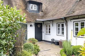 A thatched-roof cottage with a black door, white windows, and a small garden with various plants and a bench. - Haus Süderende - Gemütliches Appartement mit 34 qm Nutzfläche Unter Reet (Sylt-Ost)