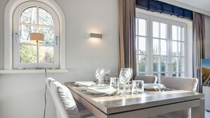 A dining room with a wooden table set for four, white upholstered chairs, a lamp, and a window with curtains. - Litz Hüs - Litz Hüs - Reetgedeckte Wohnung Liegt Idyllisch im Süden von Keitum (Sylt / OT Keitum)