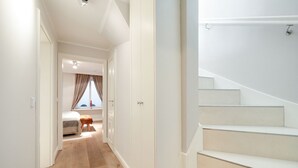 A hallway leading to a bedroom with a bed, a small stool, and a window with curtains. There is a staircase to the right. - Litz Hüs - Litz Hüs - Reetgedeckte Wohnung Liegt Idyllisch im Süden von Keitum (Sylt / OT Keitum)