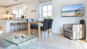 A modern kitchen with white cabinets, a dining area with a glass table, and a living room with a sofa and a TV displaying a beach scene. - Gode Tiet - Gode Tiet - Schöner Neubau im Herzen von Westerland (Sylt)