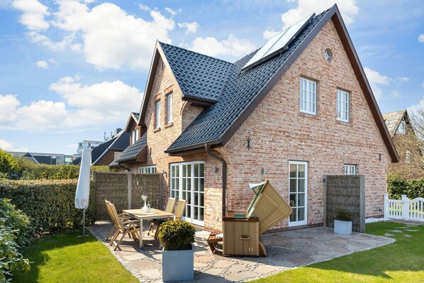 Une maison à deux étages, construite en briques, avec un toit en pente, une terrasse dotée d’une table et de chaises, et un petit jardin.