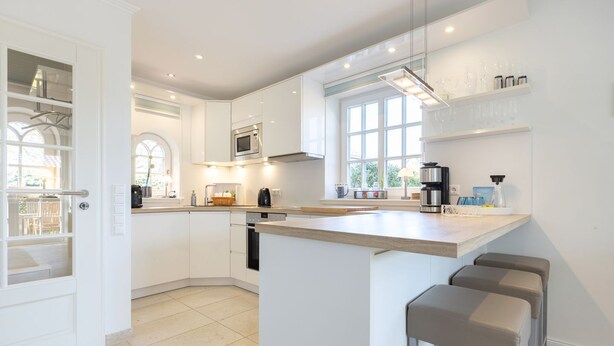 A modern kitchen with white cabinetry, a central island, bar stools, and a window providing natural light. - Gode Tiet - Gode Tiet - Schöner Neubau im Herzen von Westerland (Sylt)