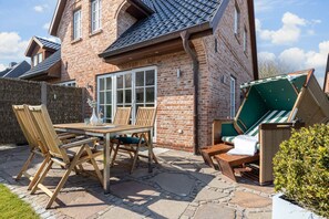 Une maison en briques avec un patio, une table en bois et deux chaises longues.