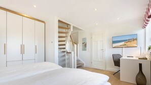 A bedroom with a large bed, a desk, and a chair, a staircase, and a door leading to another room. - Gode Tiet - Gode Tiet - Schöner Neubau im Herzen von Westerland (Sylt)