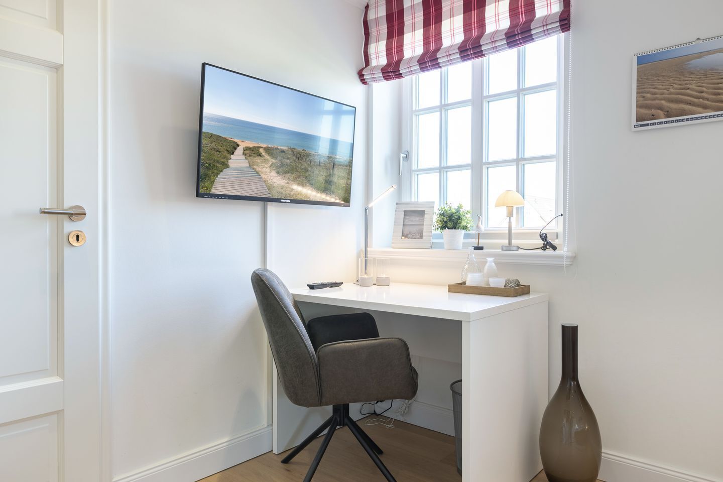 A room with a desk, a chair, a TV mounted on the wall, a window with a view of the beach, and a potted plant on the windowsill.