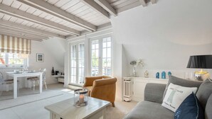 A spacious living room with a white table, a grey sofa, and a brown armchair. There is a dining area with a white table and chairs. Large windows allow natural light to fill the room. - Kleine Heimat - 2-raum-appartement im OG, ca. 50m² Nutzfläche (Sylt)