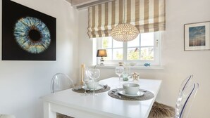 A dining area with a white table set for two, a large abstract painting, and a window with a striped valance. - Kleine Heimat - 2-raum-appartement im OG, ca. 50m² Nutzfläche (Sylt)
