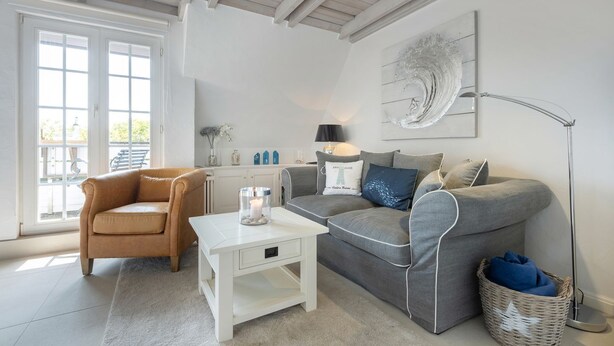 A modern living room with a beige armchair, a grey sofa, a white coffee table, and a floor lamp. There is a large window with a view of the outdoors. - Kleine Heimat - 2-raum-appartement im OG, ca. 50m² Nutzfläche (Sylt)