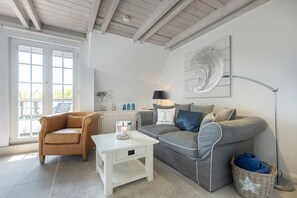 A modern living room with a beige armchair, a grey sofa, a white coffee table, and a floor lamp. There is a large window with a view of the outdoors. - Kleine Heimat - 2-raum-appartement im OG, ca. 50m² Nutzfläche (Sylt)