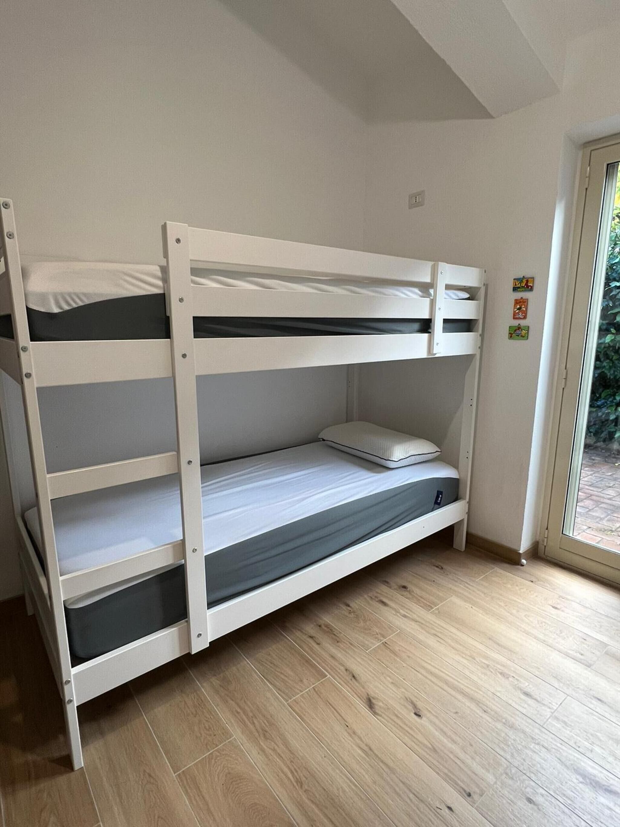 Un letto a castello con materasso e cuscini, pavimento in legno e una finestra con vista su piante.