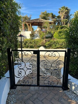 A wrought iron gate with intricate designs, leading to a paved area with a lamp post and a view of a building with a tiled roof. - Villa 'Edith' with Sea View, Shared Pool, and Air Conditioning (Parghelia)