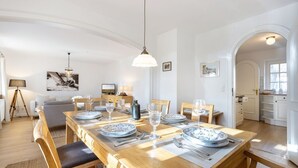 Un coin repas avec une table en bois, dressée pour quatre personnes, entourée de chaises. Un téléviseur est posé sur un meuble à gauche, et une lampe sur un trépied se trouve au premier plan. La pièce a des murs blancs et un ventilateur de plafond.