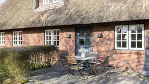 Une maison en briques avec un toit de chaume, une porte d’entrée et une terrasse avec une table et des chaises.