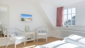Un dormitorio con una cama, dos sillones de mimbre, una mesita, un calendario, una ventana con cortinas y un radiador.
