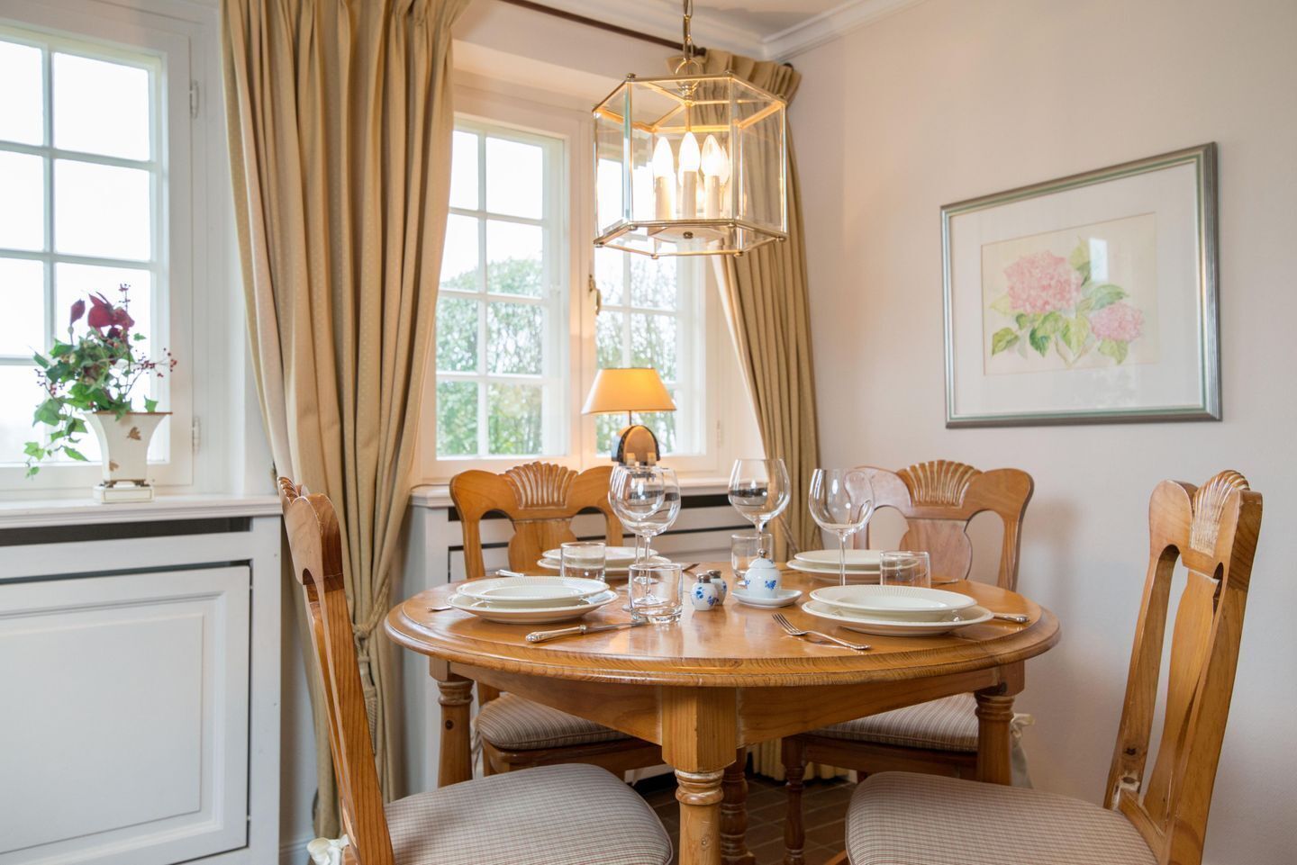 A dining room with a round wooden table set for six, surrounded by six wooden chairs with cushioned seats. A chandelier hangs above the table, and a window with curtains is visible in the background.