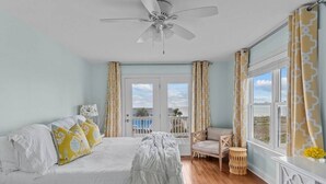 A bedroom with a bed, a ceiling fan, a window with curtains, a chair, and a small table. - Ocean View! Pool, Waterslide, Elevator & Firepit (Oak Island)