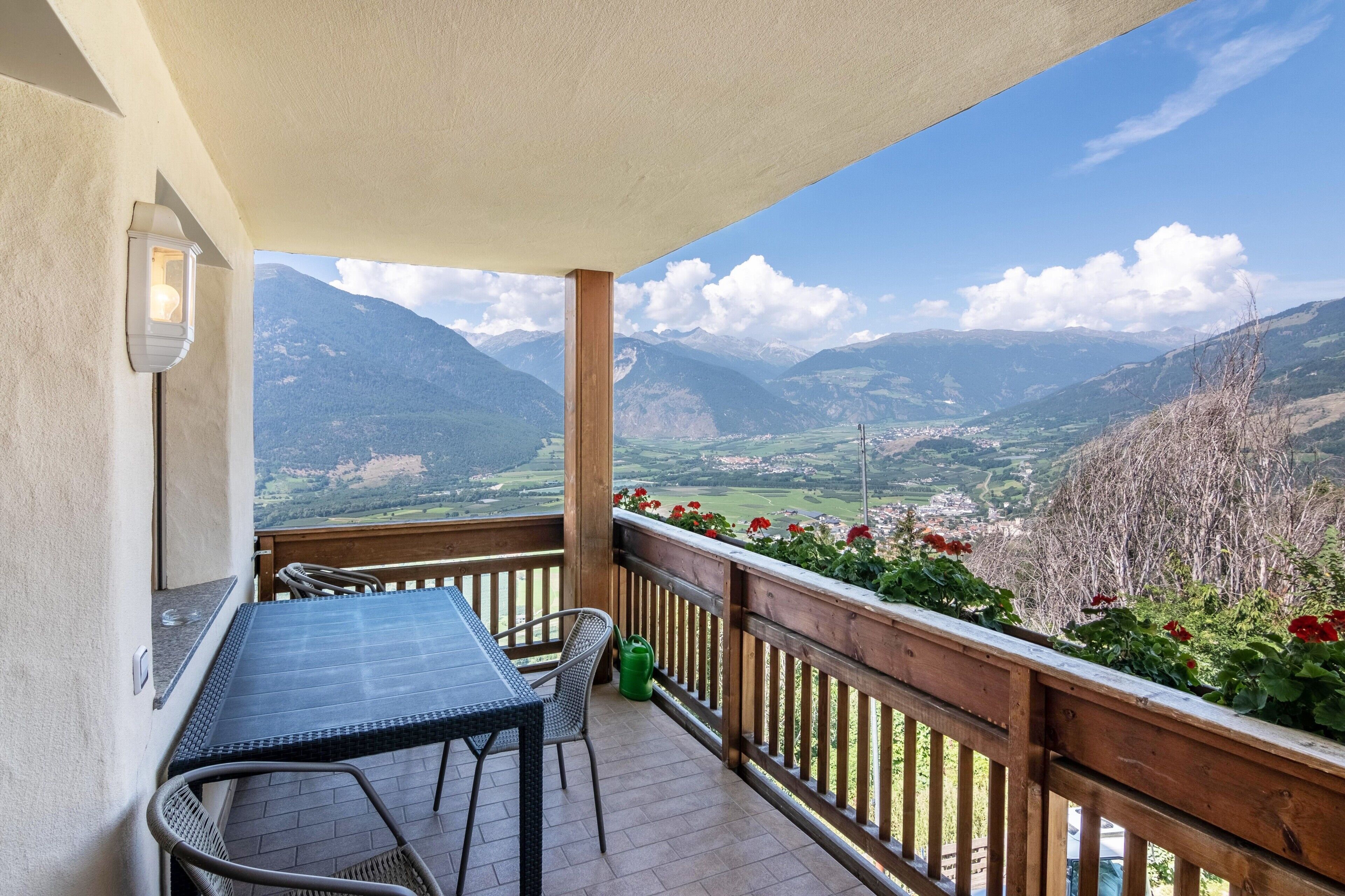 Ein Balkon mit Tisch und Stühlen, einem Hollerleger und Blick auf Berge und ein Tal.