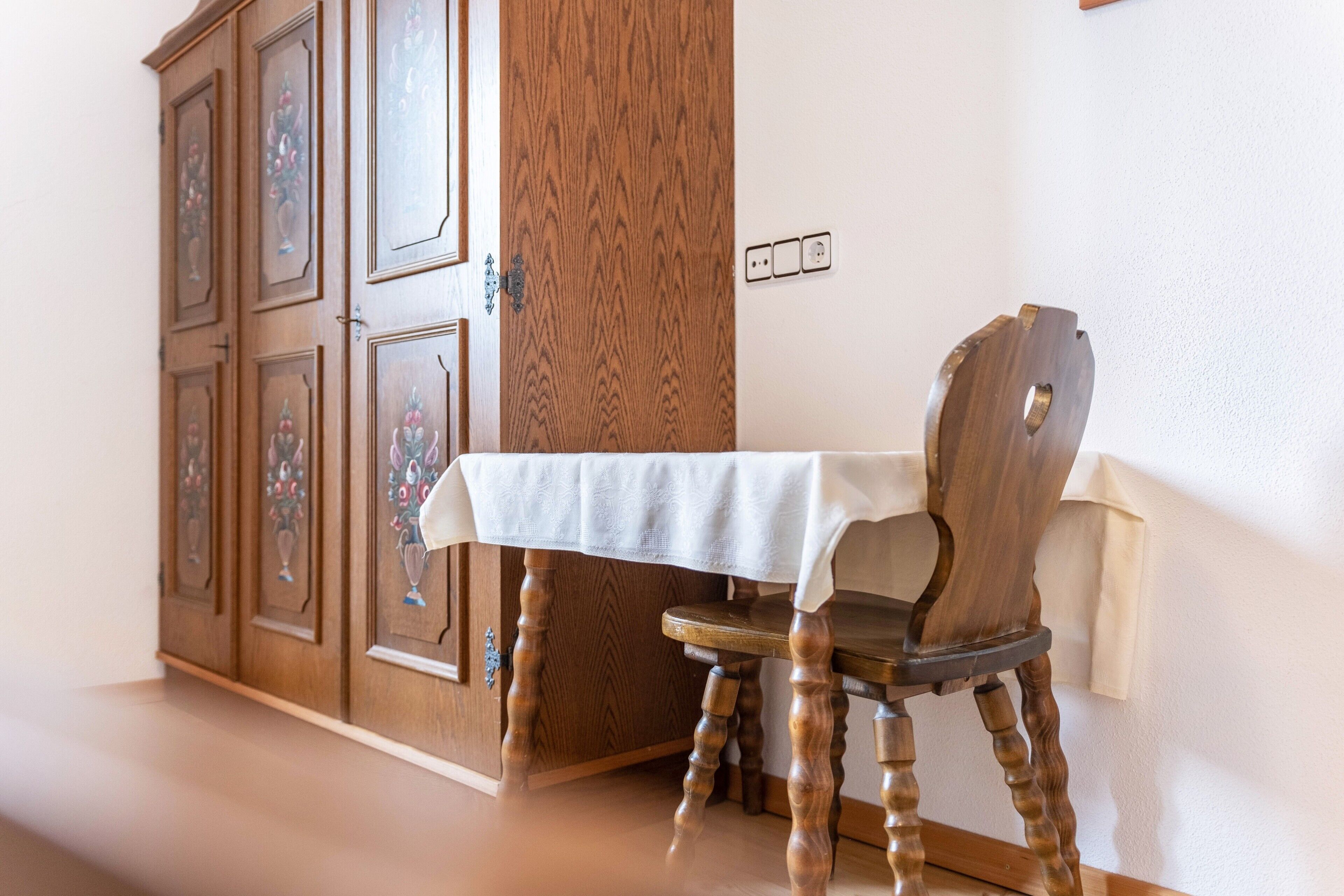 A wooden cabinet with floral designs, a table with a white tablecloth, and a wooden chair.