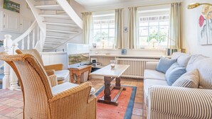 Un salón espacioso con escalera blanca, muebles de mimbre, televisor de pantalla plana y ventanas amplias con cortinas.