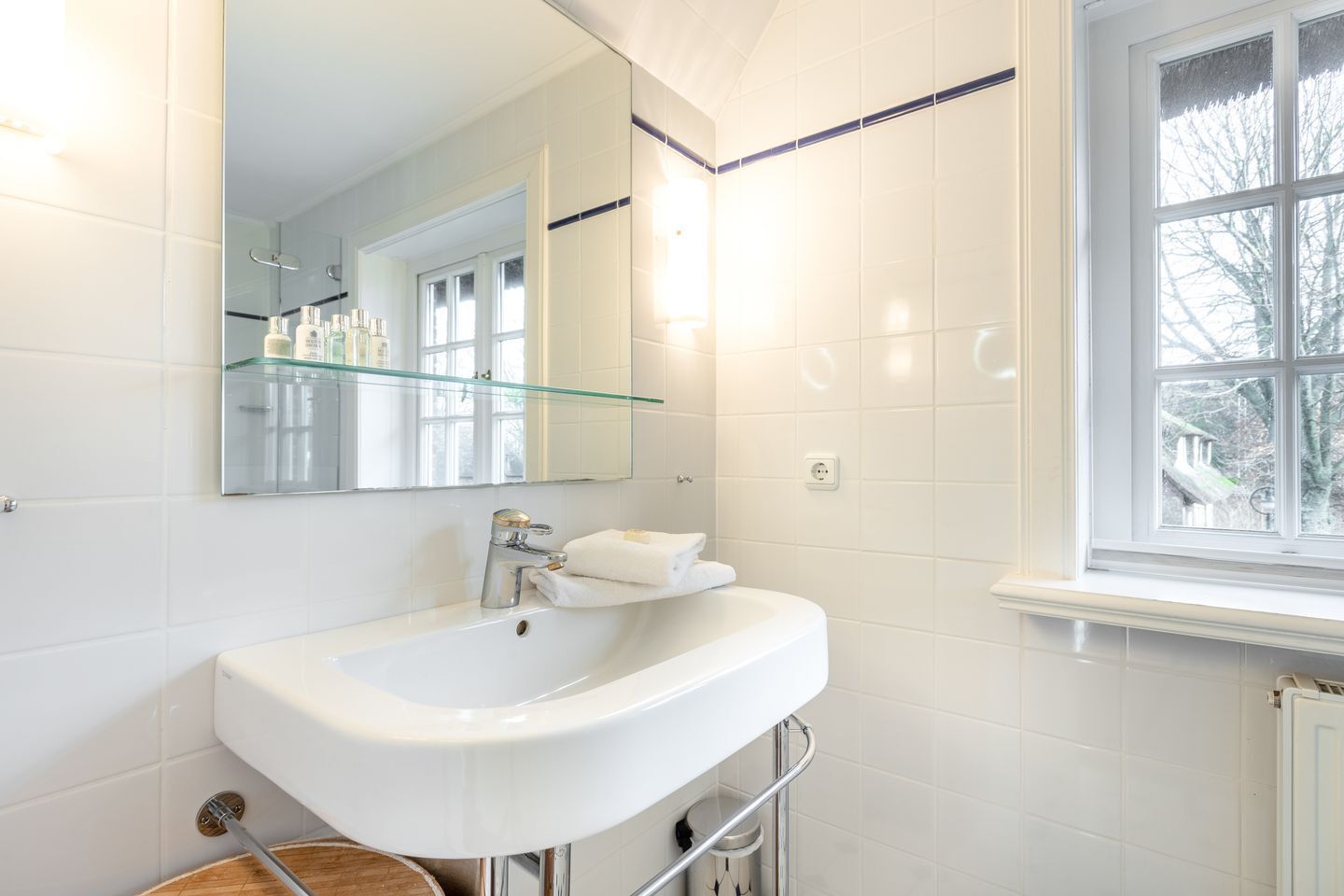 Une salle de bain moderne avec un lavabo blanc, une étagère en verre au-dessus du miroir et une fenêtre donnant sur des arbres.