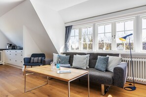 A modern living room with a sofa, a coffee table, and a dining table. There is a window with curtains and a radiator. - Blue Haven - Gemütliche Wohnung in Erster Reihe zum Wattenmeer (Sylt)