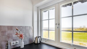 Une cuisine avec un plan de travail noir, un vase rempli de fleurs, une bouilloire et une vue sur un champ verdoyant par une grande fenêtre.