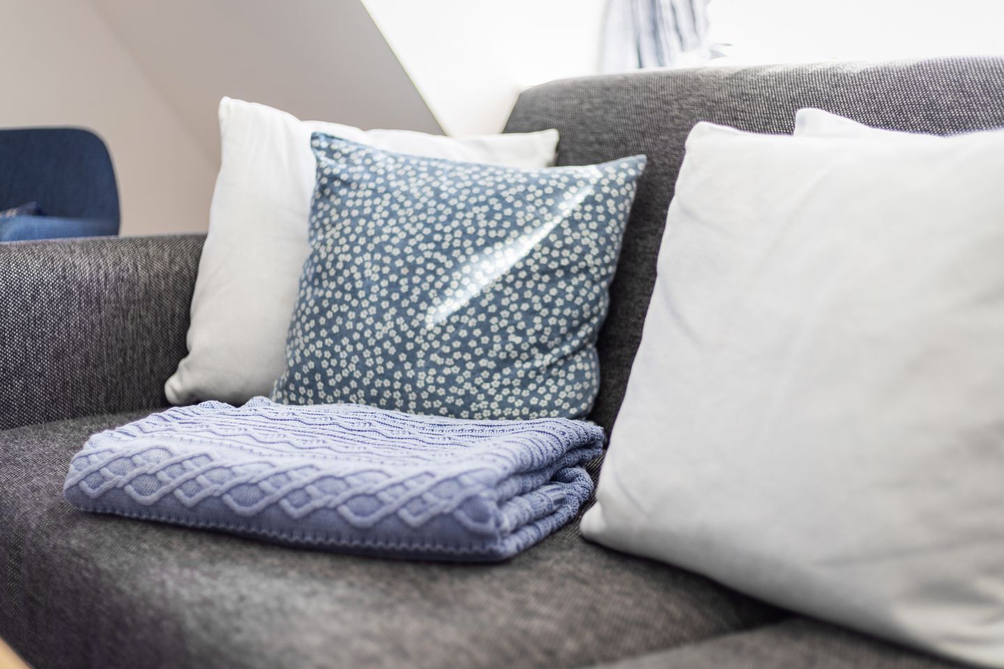 A grey sofa with three pillows: one white, one with a blue floral pattern, and one folded blue blanket.