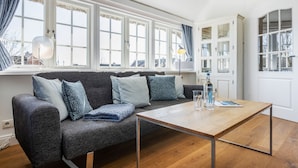 Un salon moderne avec un canapé gris, de nombreux coussins, une table basse en bois et une grande fenêtre avec des rideaux.