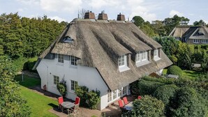 Une grande maison blanche au toit de chaume, entourée d’une végétation luxuriante et d’une pelouse bien entretenue.