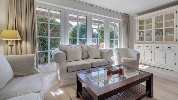 Un salon spacieux avec un canapé beige, une table basse en verre et un fauteuil blanc. De grandes fenêtres offrent une vue sur la verdure environnante.