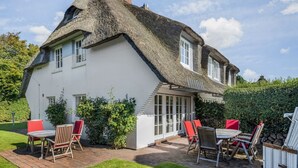 Une maison blanche avec un toit de chaume, une terrasse dotée de mobilier d’extérieur et un jardin bien entretenu.
