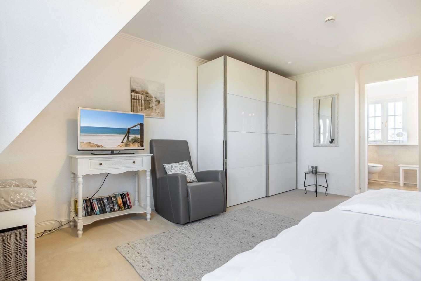 A bedroom with a bed, a TV on a white dresser, a grey armchair, and a sliding closet door.